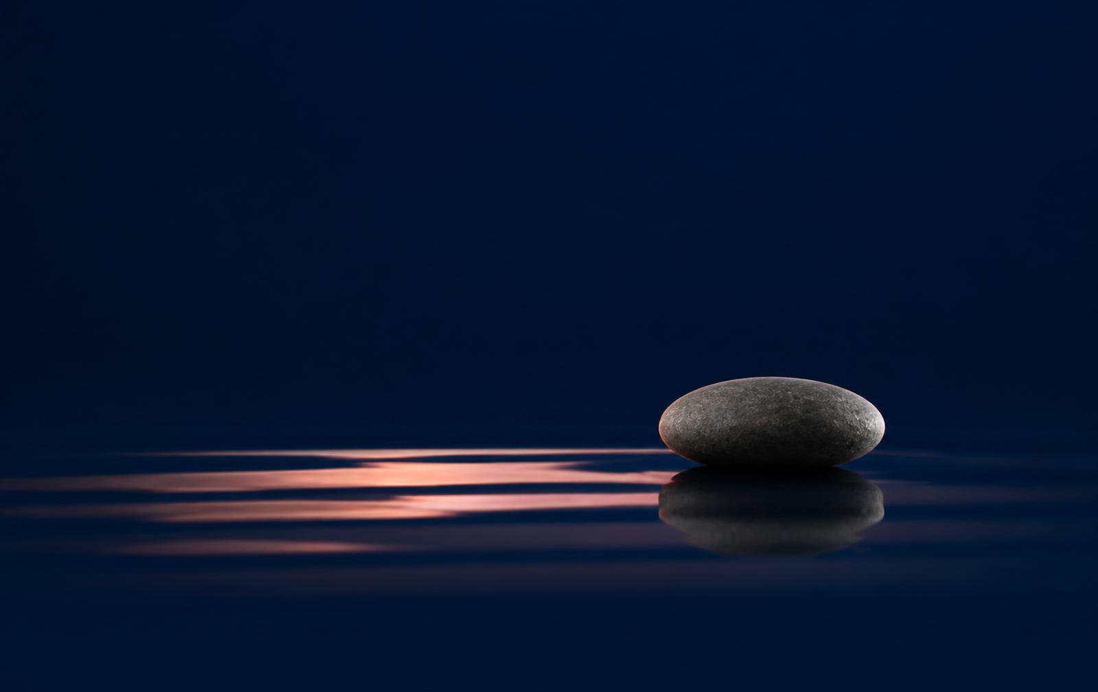 A single dark stone rests on a mirror-still water surface — a symbol of inner calm before every decision.